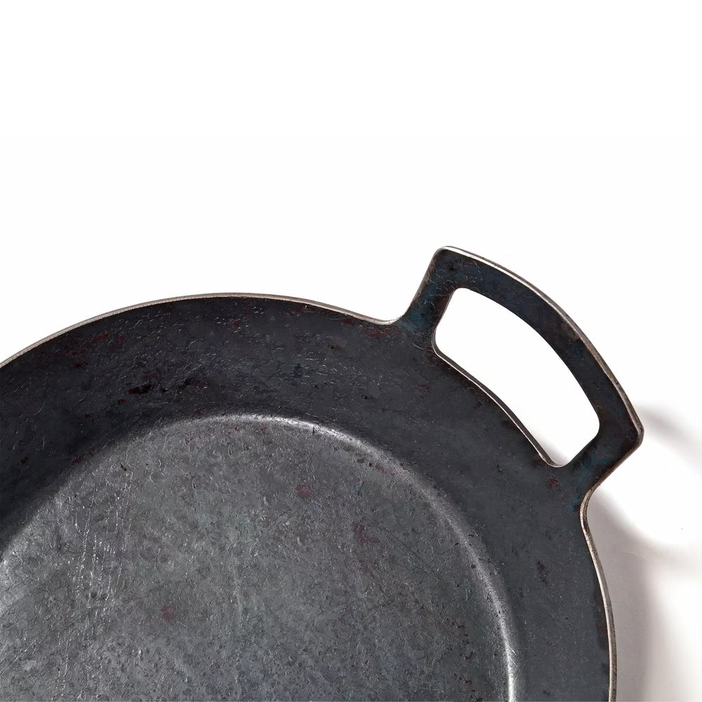 Eine Nahaufnahme der Albert Turk GmbH & Co. KG Geschmiedete Servierpfanne, hoher Rand zeigt die runde, geschwärzte Form und den Griff vor einem schlichten weißen Hintergrund; nur ein Teil der Pfanne und des Griffs sind sichtbar.