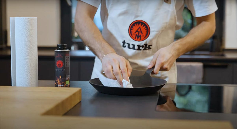 Eine Person, die eine weiße Schürze mit der Aufschrift "Turk" trägt, wischt eine schwarze Bratpfanne mit einem Papiertuch ab; eine Rolle Papiertücher und eine Flasche Öl stehen auf dem Küchentisch daneben.