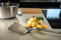 Ein Eisen-Pfannenwender von Albert Turk GmbH & Co. KG liegt mit feiner Patina auf der Arbeitsplatte neben gelben Kartoffeln und frischem Dill; im Hintergrund stehen Metallschüssel und Schneidebrett.