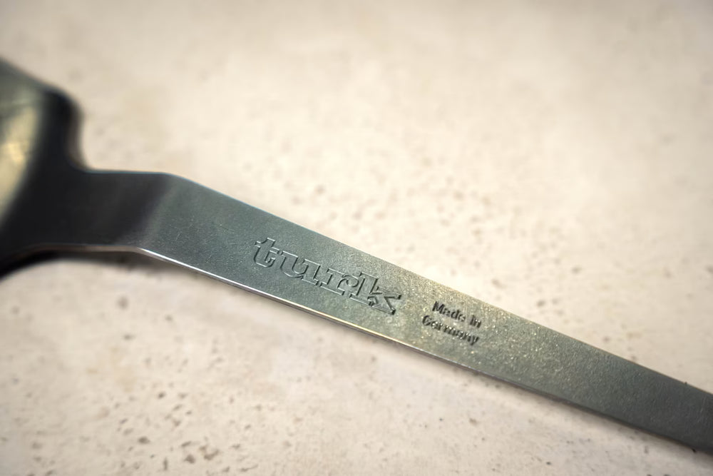 Eine Nahaufnahme eines Eisen-Pfannenwender-Griffs der Albert Turk GmbH & Co. KG zeigt die Gravur "Made in Germany" auf dem Metall, das eine dezente Patina aufweist, vor einem hell gesprenkelten Hintergrund.