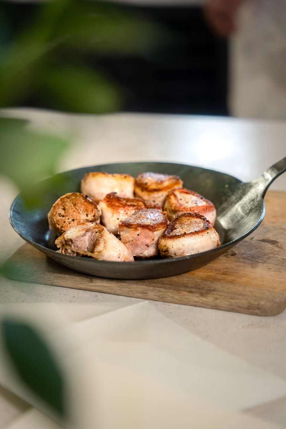 Eine Geschmiedete Bratpfanne von Albert Turk GmbH & Co. KG steht auf einem Holzbrett und enthält goldbraune Fleischmedaillons, vermutlich Schwein oder Huhn, in einer Küche mit grünen Blättern im Vordergrund.