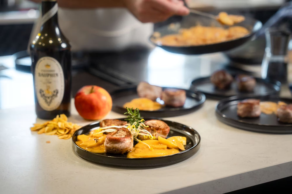 Auf einem schwarzen Teller liegen gebratene Fleischmedaillons auf gelber Mango, garniert mit Kräutern. Im Hintergrund kocht eine Person mit einer Albert Turk GmbH & Co. KG Geschmiedete Bratpfanne auf einer weißen Arbeitsplatte, auf der eine Flasche, ein Apfel und weitere Teller zu sehen sind.