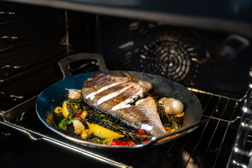 Ein ganzer Fisch mit geritzter Haut brät in einer Albert Turk GmbH & Co. KG Geschmiedete Servierpfanne, umgeben von Kräutern, Knoblauch, Zitrone und Chilischeiben in einer schlichten schwarzen Pfanne.