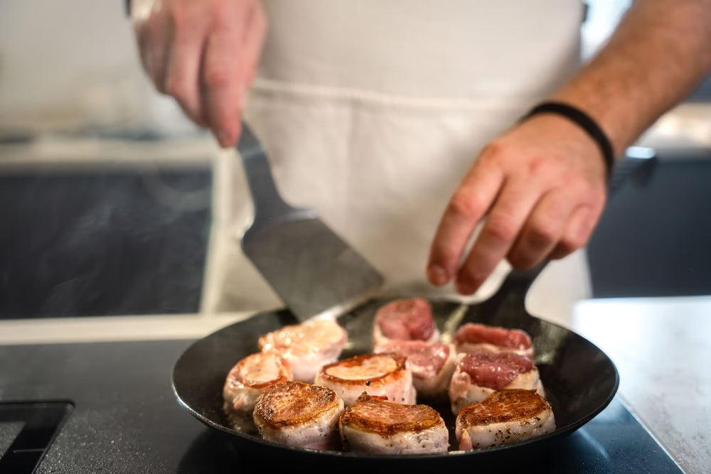 Eine Person in weißer Schürze brät mit einem Pfannenwender rundes, gewürztes Fleisch in einer Albert Turk GmbH & Co. KG Geschmiedete Bratpfanne auf einer Herdplatte.