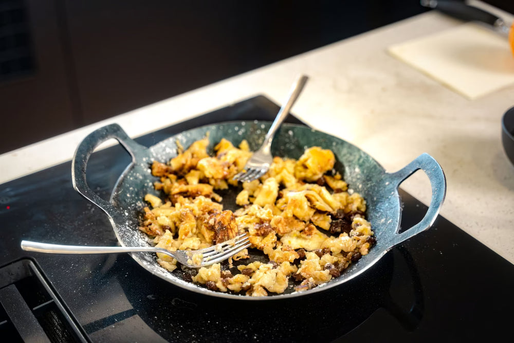Eine Geschmiedete Servierpfanne von Albert Turk GmbH & Co. KG mit gebräunten Rühreiern steht auf der Herdplatte. Zwei Gabeln liegen darin, bereit zum Teilen. Im Hintergrund sind die Küchentheke und das Schneidebrett zu sehen.