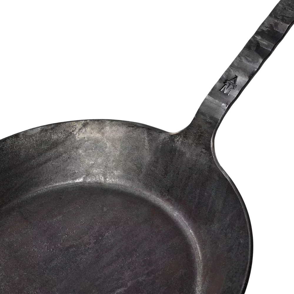 Eine Nahaufnahme der Geschmiedeten Bratpfanne der Albert Turk GmbH & Co. KG zeigt die schwarze, gewürzte Gusseisenoberfläche und den Metallgriff mit eingeprägtem Pfeildesign.